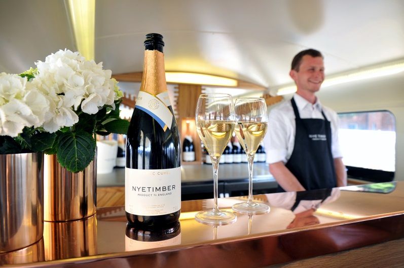 a wine bottle, two glasses and a bouquet of white flowers on a bar, waiter in the background