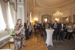 a woman holding a glass of wine, speaking at an event to a room of spectators