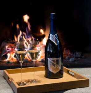 a bottle of Nyetimber 1086 Prestige Cuvee wine and two wine glasses on a wooden tray in front of a burning fire